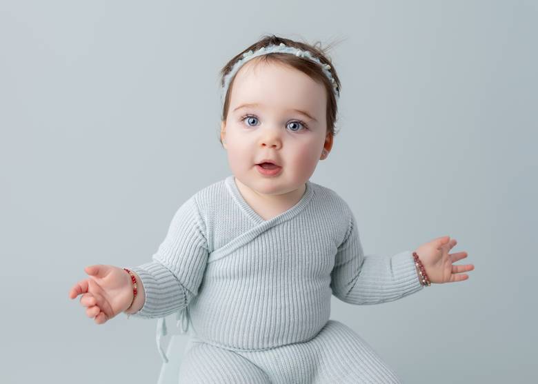 Baby in a pale green knit set with both hands open, mid babble.