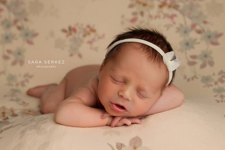 Newborn asleep on a soft floral backdrop wearing a cream headband.