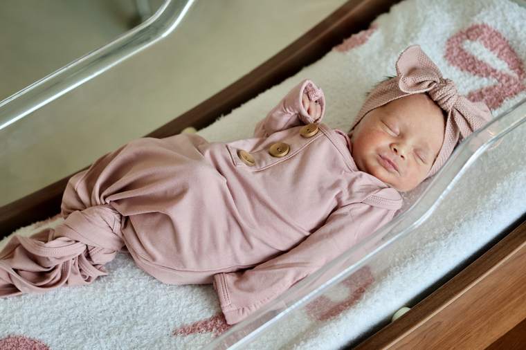 Newborn in a pink outfit and bow asleep in a hospital bassinet.