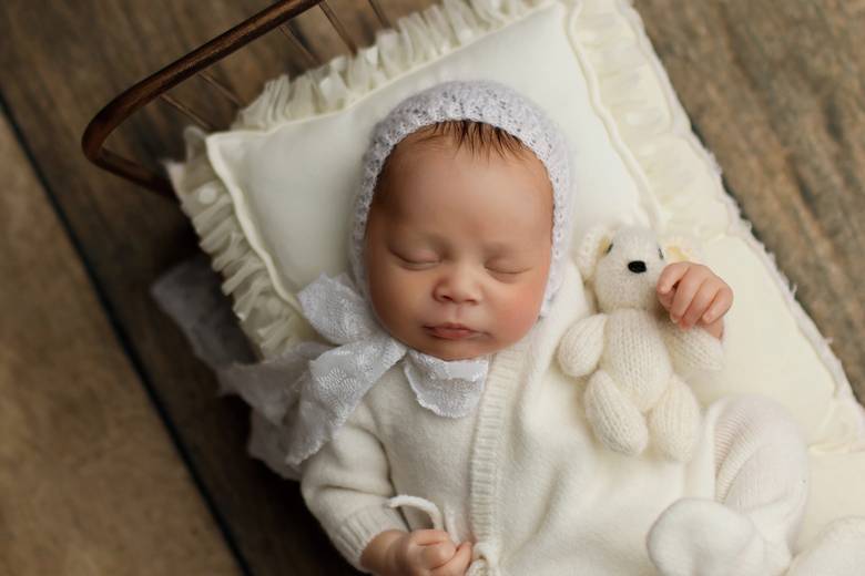 Newborn in a white bonnet asleep beside a small knitted bear.