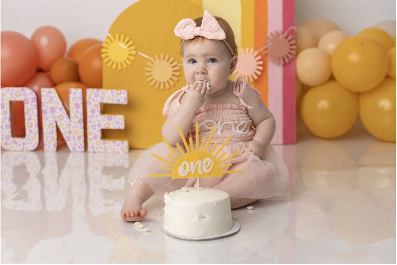One year old in a pink dress eating cake at a sunny cake smash session.