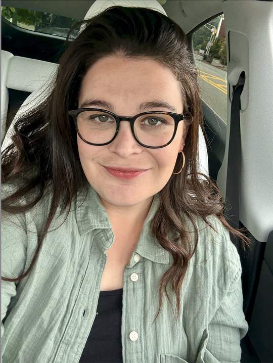 Sara Serkez, dark hair and tortoiseshell glasses, smiling in her car.