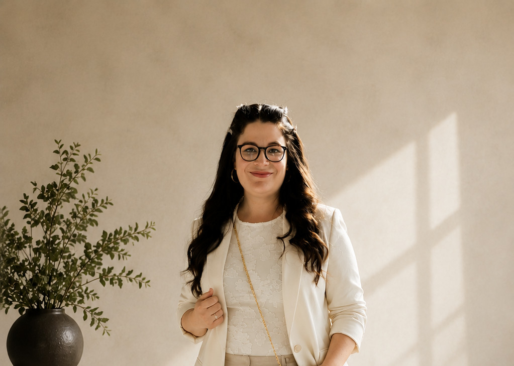 Sara Serkez in a cream blazer, standing in warm window light beside a potted plant.