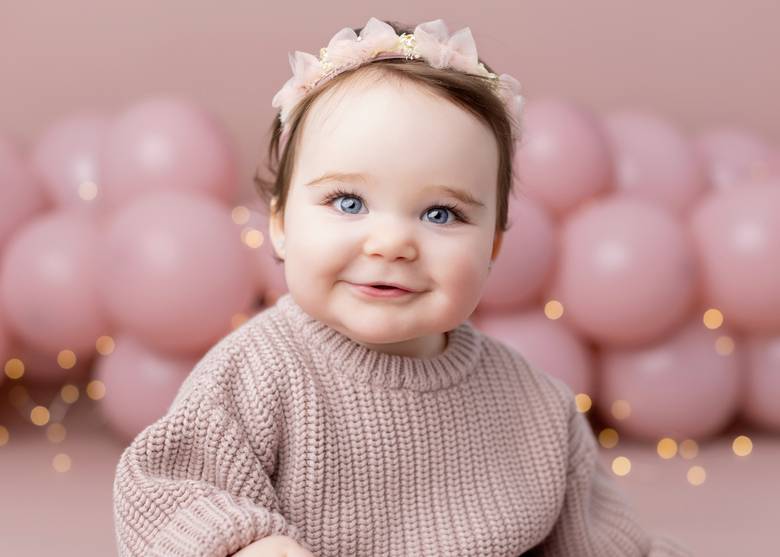 Smiling one year old in a pink knit sweater against a pink balloon backdrop.